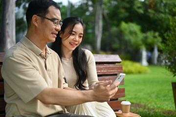 Happy middle age man and adult daughter watching funny video on smart phone while sitting on bench...