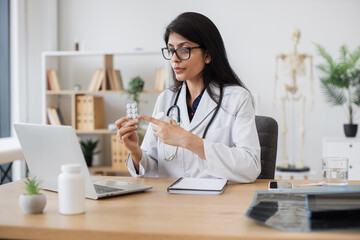 Attentive indian woman holding medicines and telling about it effects during video call on wireless laptop. General practitioner in medical uniform having online appointment with sick patient.