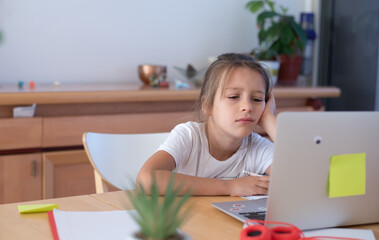 Child study at home. Girl in white t-shirt sit in table look on computer. study on zoom online. Kid hold the hand on head with thoughtful face task exersice
