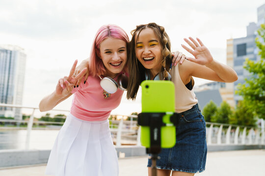 Two smiling girls recording videos with smartphone for their social media vlog on city waterfront