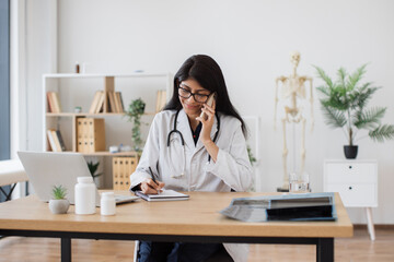 Busy female doctor speaking on modern smartphone and writing patient problems in notebook. Focused indian woman with stethoscope on neck sitting at table and listening complaints about health.