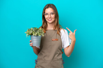 Young caucasian gardener woman holding a plant isolated on blue background giving a thumbs up gesture