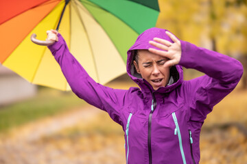 A young woman with an umbrella in a rain jacket wears a hood and protects her head from rain and strong wind during late autumn