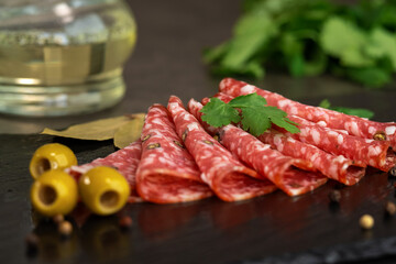 Fresh slices smoked sausage with spices and seasonings on stone board with olives on background of greenery. Cold cuts. Meat. Food