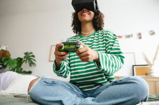 Happy girl wearing virtual reality simulators playing video game with controller in bedroom