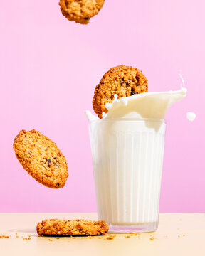 Flying oatmeal cookies near milk splashing in glass against pink colored background