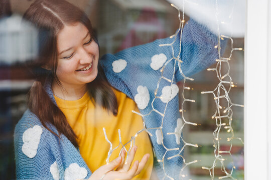 Happy teenage girl decorating with string lights seen through glass