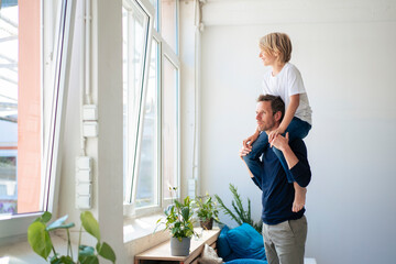 Thoughtful father carrying son on shoulders and looking through window at home