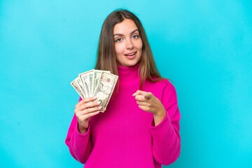 Young caucasian woman taking a lot of money isolated on blue background surprised and pointing front