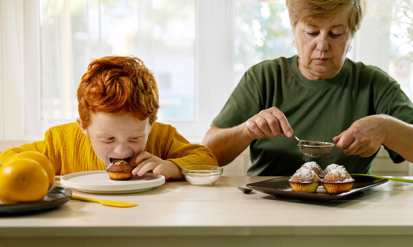 Grandmother dusting sugar on cupcakes with grandson eating in kitchen at home - Powered by Adobe