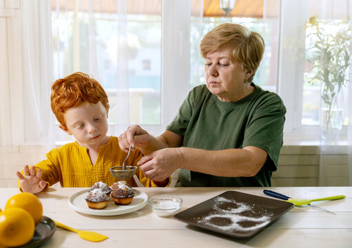 Grandmother sprinkling sugar on cupcakes for grandson at kitchen