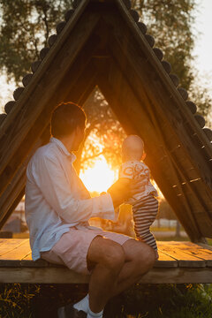 Father and baby boy sitting in wooden cabin and watching sunset at park