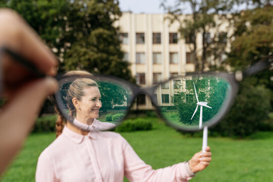 Smiling businesswoman holding wind turbine model seen through concave eyeglasses at park