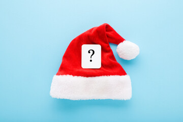 White card of black question mark on bright red Santa Claus hat on light blue table background. Pastel color. Closeup. Christmas issues concept. Top down view.