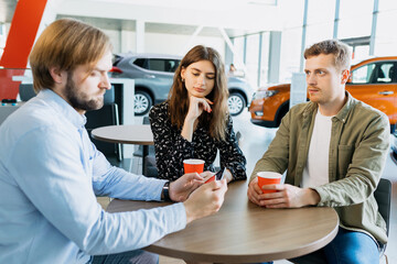 A young couple is sitting at a table with a sales manager who shows on a tablet which cars can be bought at a dealership. The long-awaited purchase of a family car.