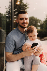 Fototapeta premium Young man using a phone mobile for work and holding his son on his hand on the cafe. Being a daddy and working everywhere. Freelancing and networking concept