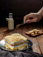 Layered cheese cake with grated cheese topping is neatly presented on a plate with a hand about to cut it, isolated against a wooden table background pattern.