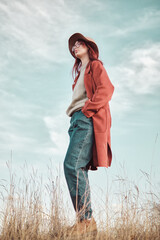 Confident teenage girl in red coat and hat standing against the sky with hands in pockets and looking away. Low angle view. © TanyaJoy