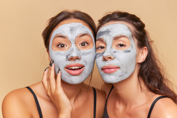 Two cute girls make cosmetic masks on their faces, look at the camera with interest, each has mask of medicinal clay on her face, skincare products concept, copy space