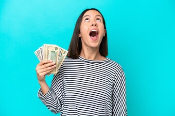 Young caucasian woman taking a lot of money isolated on blue background looking up and with surprised expression