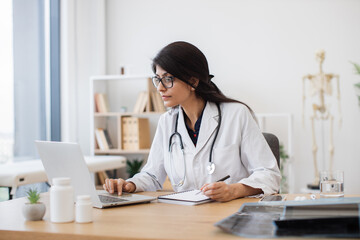 Concentrated female doctor with stethoscope sitting at table and writing prescription while using...