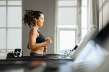 Muscular fitness woman listening to music on wireless earphones while running on a treadmill, doing exercises in the morning at a gym. 