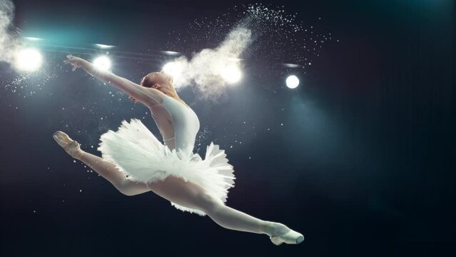 Beautiful Ballet Performance on a Big Stage with Lights. Young Female Ballerina in White Dress Making a Perfect Split Leap and Releasing Powder. Super Slow Motion Speed Ramp Effect