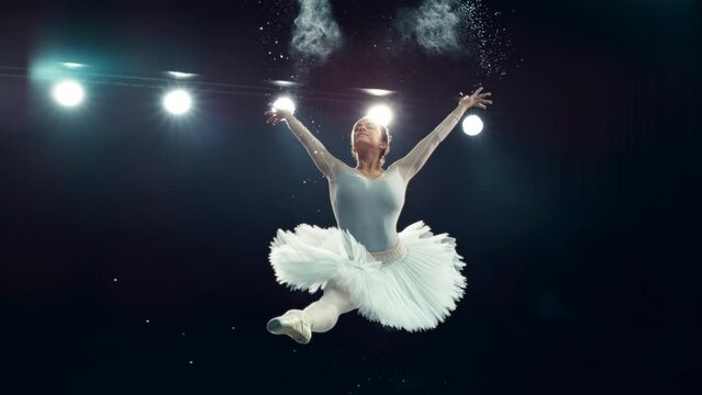 Modern Ballet Performance on a Big Dark Stage with Lights. Young Female Dancer in White Dress Making a Striking Split Leap and Releasing Powder. Super Slow Motion Speed Ramp Effect