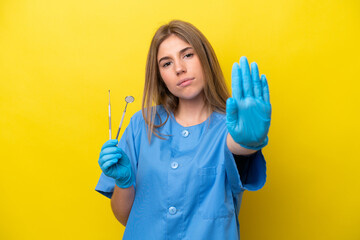 Dentist caucasian woman holding tools isolated on yellow background making stop gesture