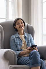 Vertical portrait of attractive middle-aged woman pose for camera sit on armchair with cellphone in hands. Modern application user, remote communication through messenger, mobile connection, lifestyle