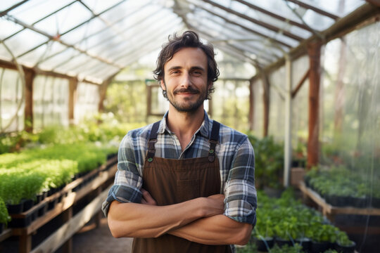 Portrait of Self assured adult man in checkered shirt and apron crossing arms and looking at camera with smile while standing on blurred background of greenhouse on farm