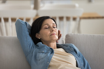Peaceful middle-aged female falling asleep on sofa at home, leaned on couch, sleeping, take healthy...