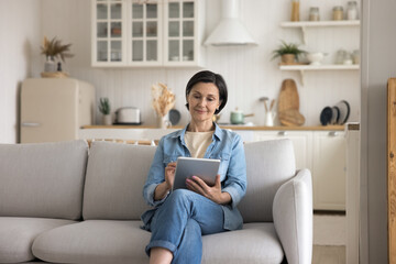 Pretty middle-aged woman spending leisure on internet sit on couch using digital tablet, choose goods, buy e-services through e-commerce app or website, enjoy easy remote electronic shopping at home