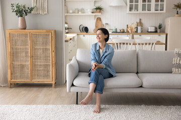 Satisfied retired pretty woman resting on sofa in modern living room, laugh, staring into distance...