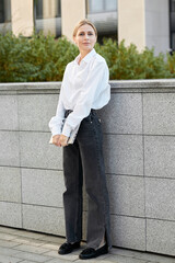 A well-groomed blond woman in a white blouse and black jeans walks around the city with a laptop,...