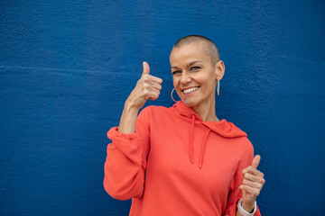 Woman with shaved head showing thumbs up in front of blue wall