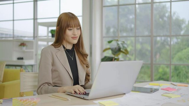 Young asian business woman smiling and typing on laptop computer working in internet, beautiful female professional user lady using pc technology doing online job in office or browsing web in office