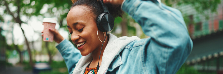Smiling African girl with ponytail wearing denim jacket, in crop top with national pattern walking down the street listening to music on headphones and dancing