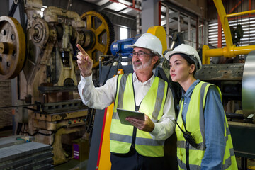 Senior engineer manager trains new employees within the metal sheet factory. Everyone wear safety vest and hardhat. Various kind of industrial machine are in the working area.