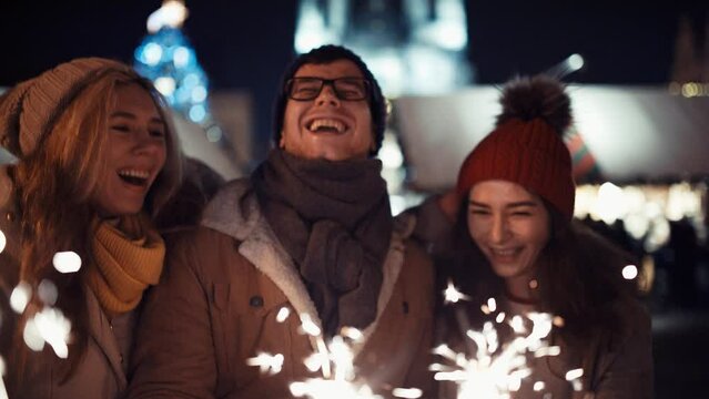 Close Up Portarait of Two girls kiss a happy guy with glasses on new year's eve to the sound of chimes in the middle of the street in the lights of the Christmas market on the background of a