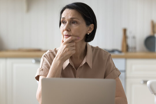 Brooding Woman Sit At Table With Laptop In Kitchen Deep In Thought, Looks Aside, Ponder On Problem, Experiences Lack Of Understanding, Thinks On Unpleasant News, Distracted From Work. Tech, Complexity