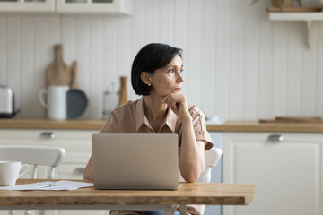 Serious middle-aged woman sit at table in kitchen with laptop, working, managing family budget,...