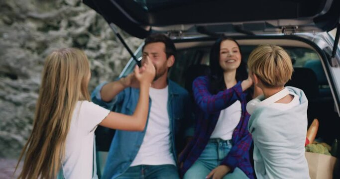 Parents man and woman sitting in car trunk at camping their smiling children girl and boy come up and give high fives and hugging. Time to Travel concept. Family travel dream concept.