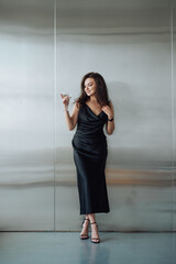 Positive stylish lady in a black dress standing with a glass of alcohol in her hand on a silver background and looking away with a smile on her face.