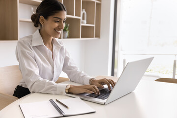 Happy beautiful young Indian business professional woman enjoying online communication at laptop, working at office table, typing on computer, laughing, smiling at screen