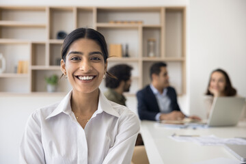 Happy young beautiful Indian businesswoman office portrait. Positive pretty business professional girl looking at camera, posing in co-working meeting room with colleagues talking behind