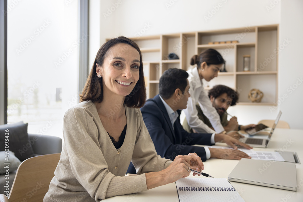 Wall mural Happy mature Latin businesswoman, project manager, female team leader portrait. Positive business woman smiling at camera while with multiethnic colleagues working at laptop in background