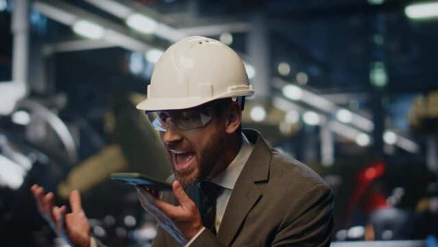 Furious boss shouting smartphone at production workshop in helmet close up.