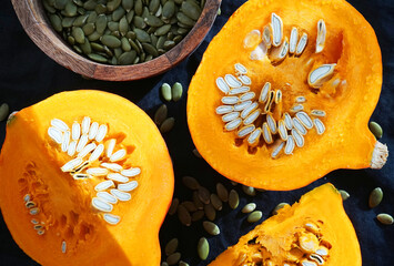 Cut pumpkin next to pumpkin seeds in a wooden plate on a dark background
