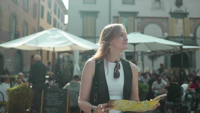 Attractive young female tourist is exploring new city. Portrait of 30s traveler woman tourist with map walking at street in Bergamo old town. Traveling Europe in autumn. 